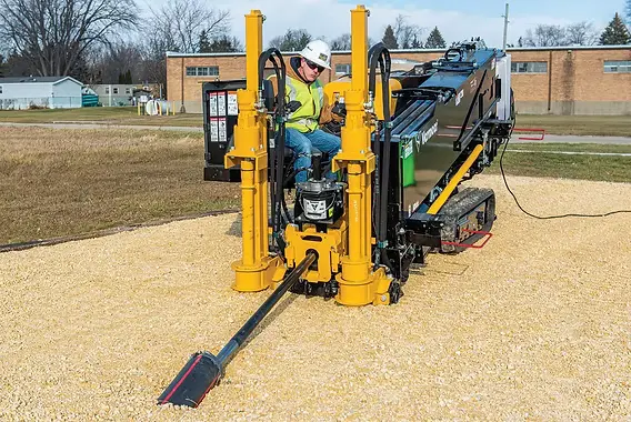 A contractor in a hard hat and high-visibility vest operating a compact horizontal directional drilling rig on a gravel pad at a fiber job site.